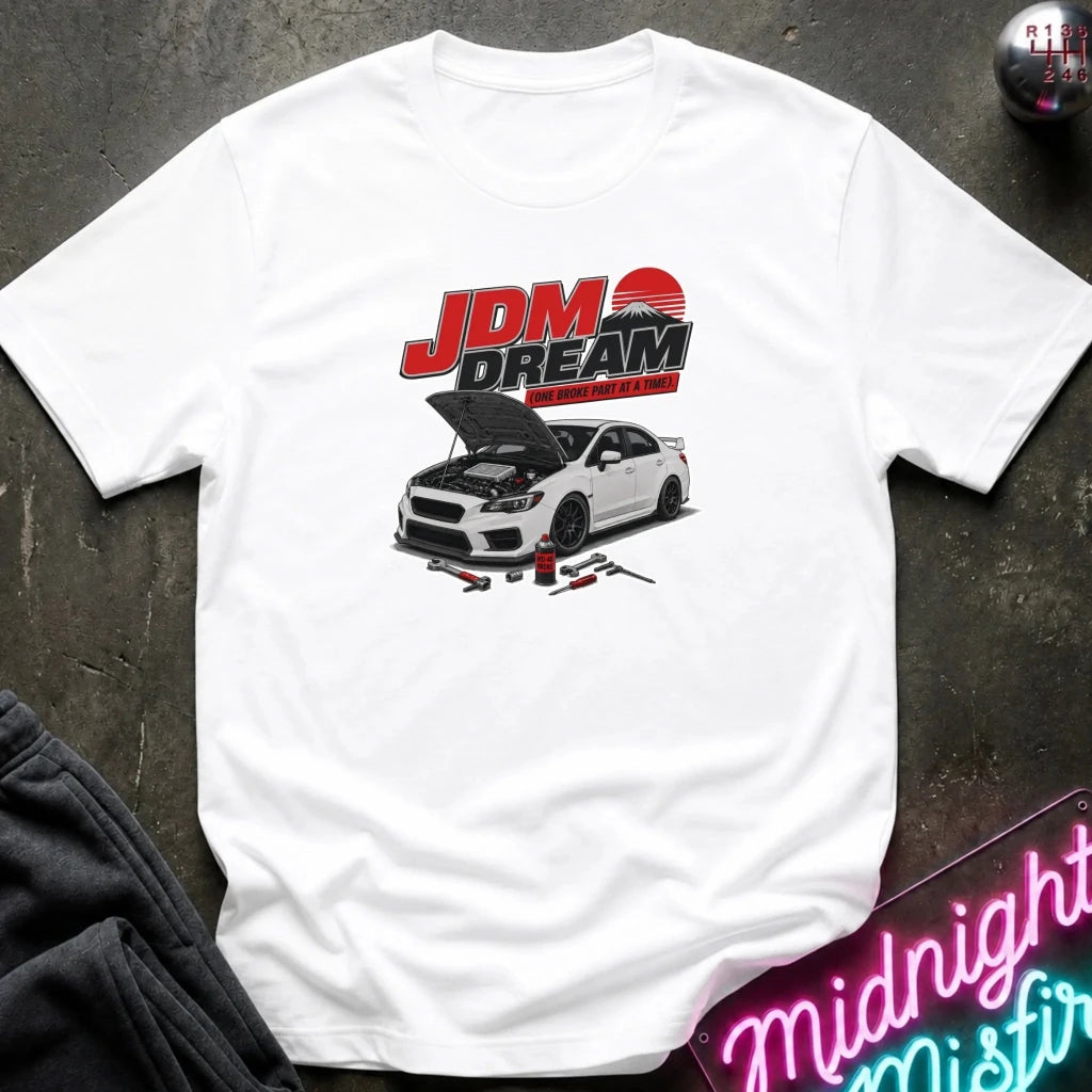 white short-sleeve graphic T-shirt laid flat on a dark textured surface. The shirt features a large front print with the text 'JDM DREAM' in red and black above an illustration of a white sports car with its hood open, plus small automotive tools.