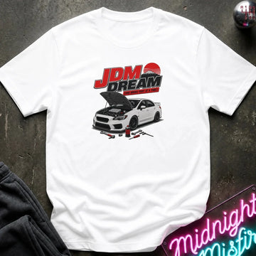 white short-sleeve graphic T-shirt laid flat on a dark textured surface. The shirt features a large front print with the text 'JDM DREAM' in red and black above an illustration of a white sports car with its hood open, plus small automotive tools.