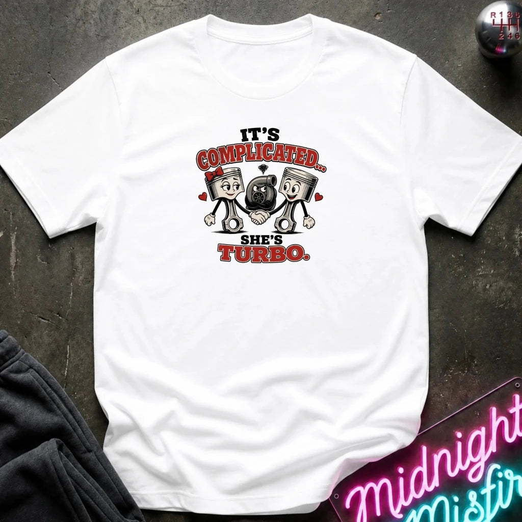 A white short-sleeve graphic T-shirt laid flat on a dark textured surface. The shirt features a cartoon graphic of two pistons holding a heart-shaped turbocharger, with the text 'IT'S COMPLICATED.