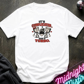 A white short-sleeve graphic T-shirt laid flat on a dark textured surface. The shirt features a cartoon graphic of two pistons holding a heart-shaped turbocharger, with the text 'IT'S COMPLICATED.