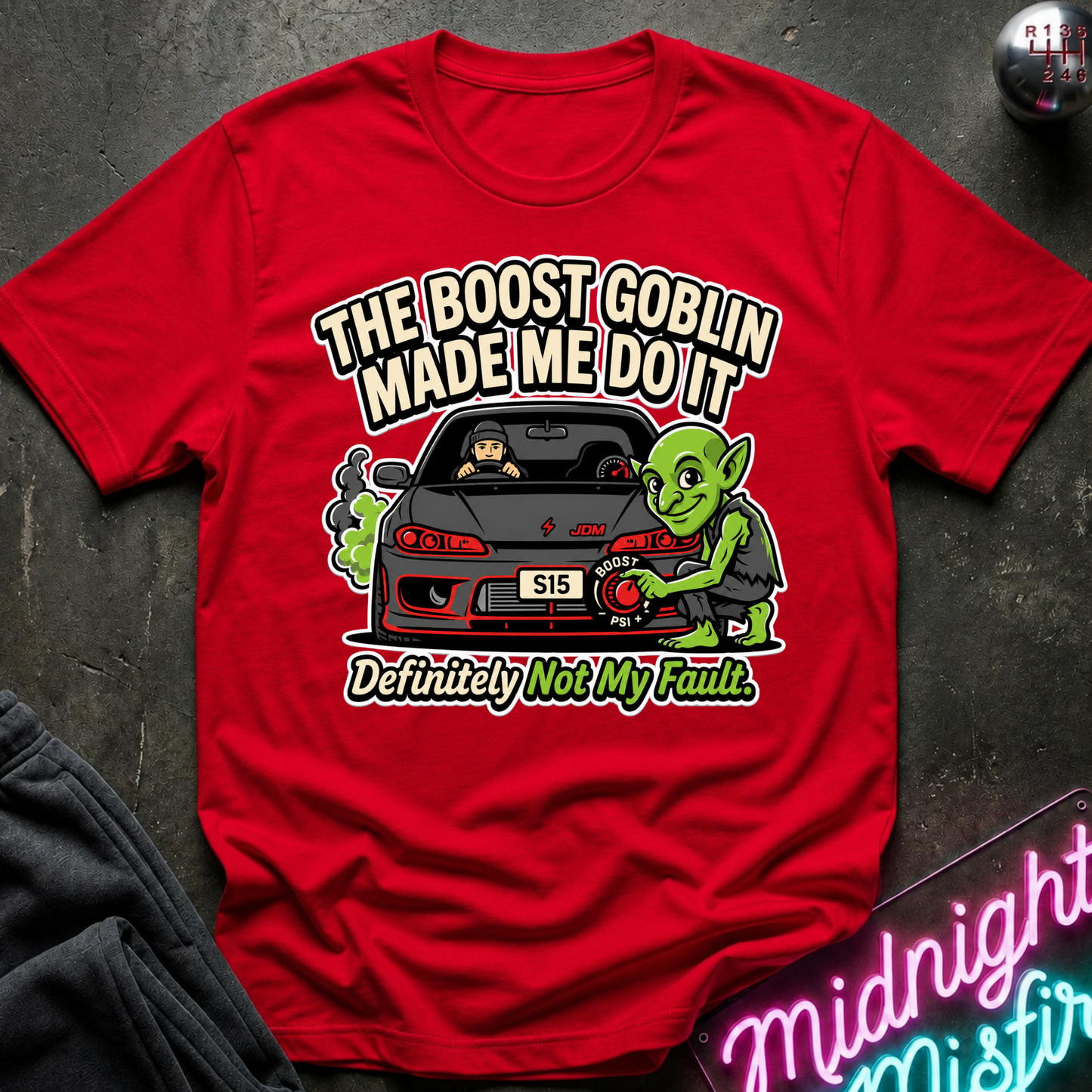 A bright red graphic t-shirt laid flat on a dark textured surface. The shirt features a cartoon illustration of a green goblin working on a black sports car, with the text 'THE BOOST GOBLIN MADE ME DO IT' above and 'Definitely Not My Fault' below.