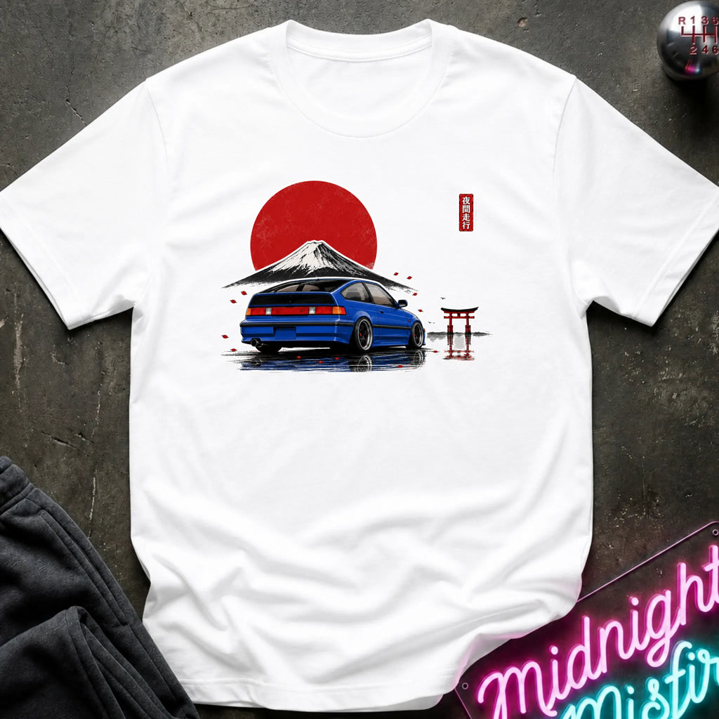A white short-sleeve t-shirt laid flat on a dark textured surface, featuring a graphic print of a blue sports car in front of a red sun and mountain, with a small red torii gate and Japanese characters. A neon sign and dark clothing are partially visible in the corners.