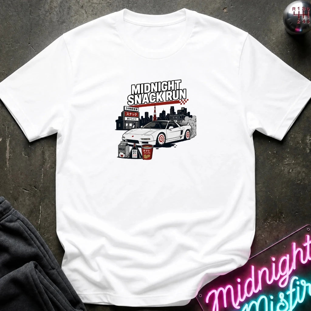 A white short-sleeve graphic t-shirt laid flat on a dark textured surface. The graphic features a white sports car with red wheels and black text reading 'MIDNIGHT SNACKIRUN' above it.