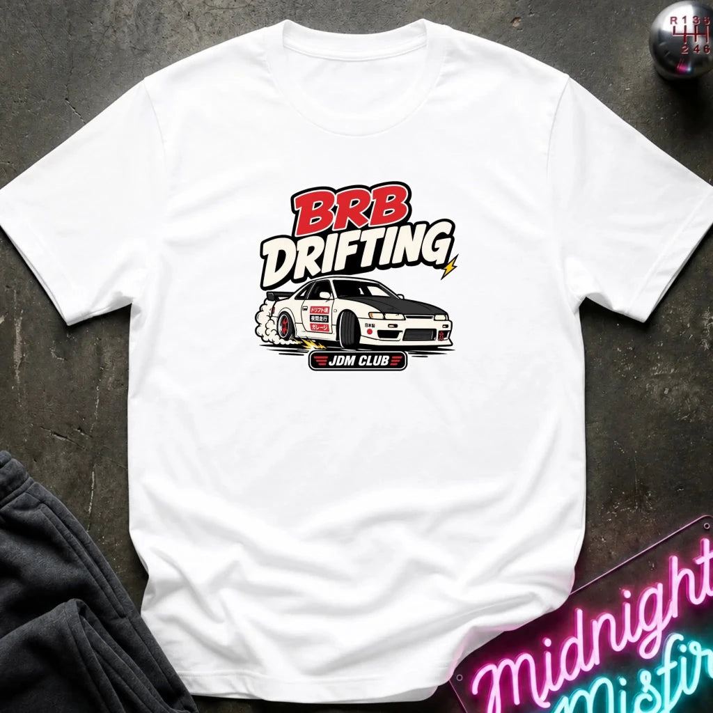 A white short-sleeve graphic T-shirt laid flat on a dark textured surface. The shirt features a 'BRB DRIFTING' 