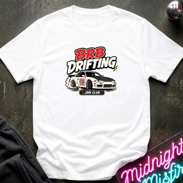 A white short-sleeve graphic T-shirt laid flat on a dark textured surface. The shirt features a 'BRB DRIFTING' 