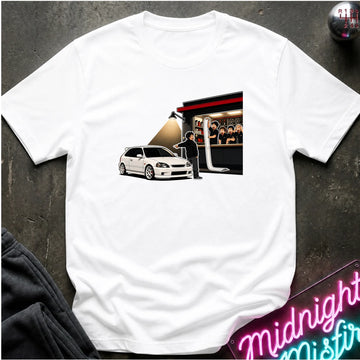 A white short-sleeve crewneck t-shirt laid flat on a dark textured surface. The front of the shirt features a rectangular graphic print depicting a white hatchback car, a person standing, and a building with people inside.