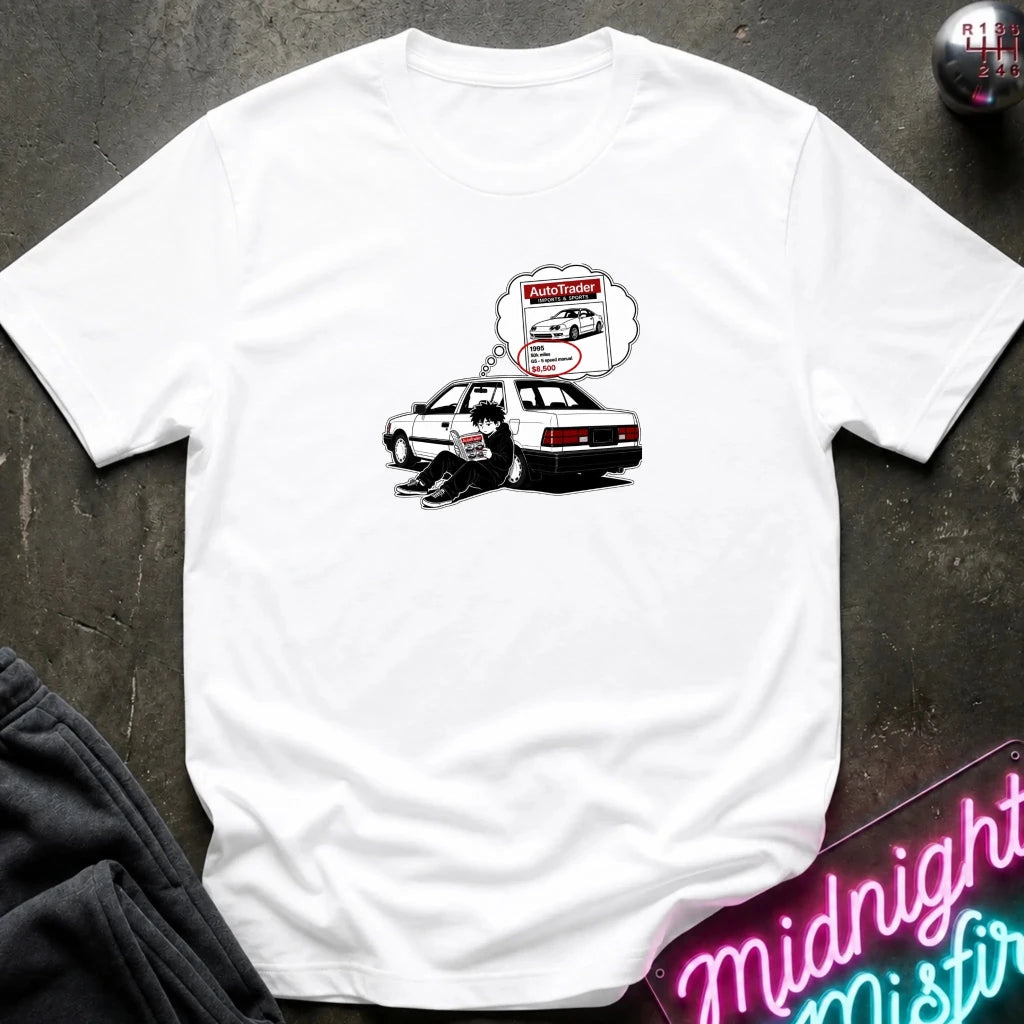 A white short-sleeve t-shirt laid flat on a dark textured surface, featuring a graphic of a black-and-whitecar with a thought bubble showing a car part diagram.
