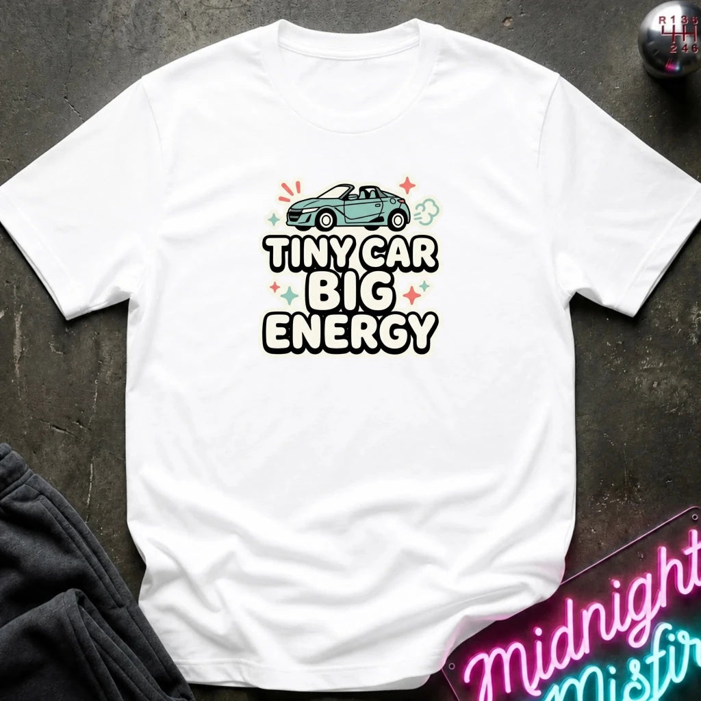 A white short-sleeve graphic T-shirt laid flat on a dark textured surface. The shirt features a teal convertible car above the bold text 'TINYCAR BIG ENERGY' with small colorful star and cloud accents.