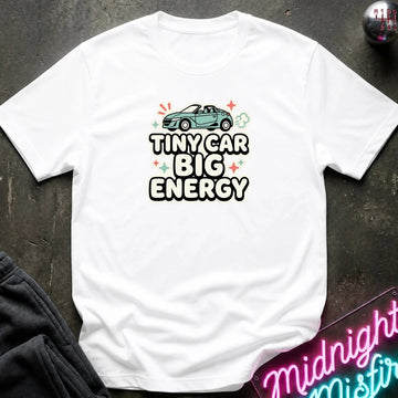 A white short-sleeve graphic T-shirt laid flat on a dark textured surface. The shirt features a teal convertible car above the bold text 'TINYCAR BIG ENERGY' with small colorful star and cloud accents.