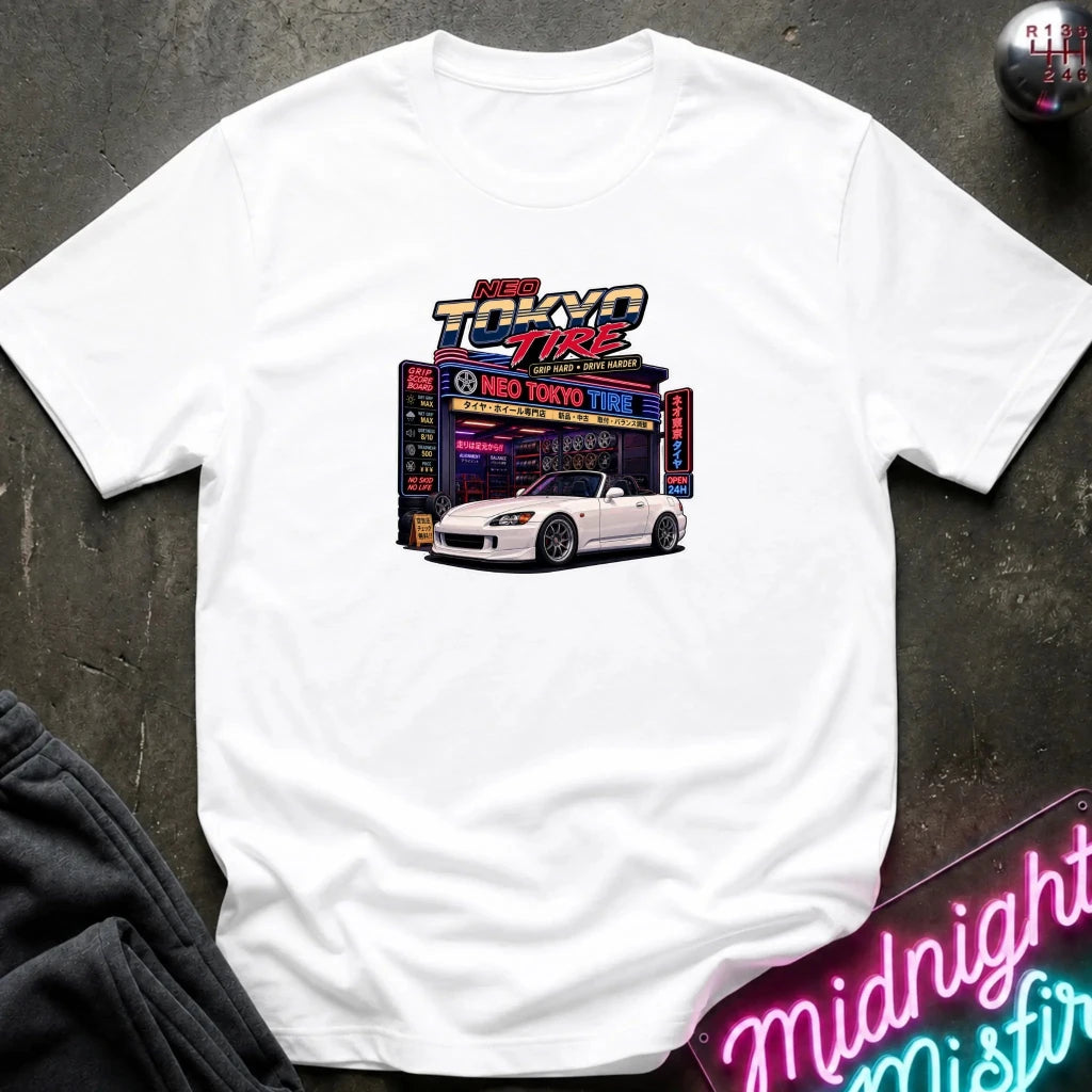 A white short-sleeve t-shirt laid flat on a dark textured surface, featuring a colorful retro graphic of a white sports car parked in front of a neon 'Neo Tokyo Tire' shop. 