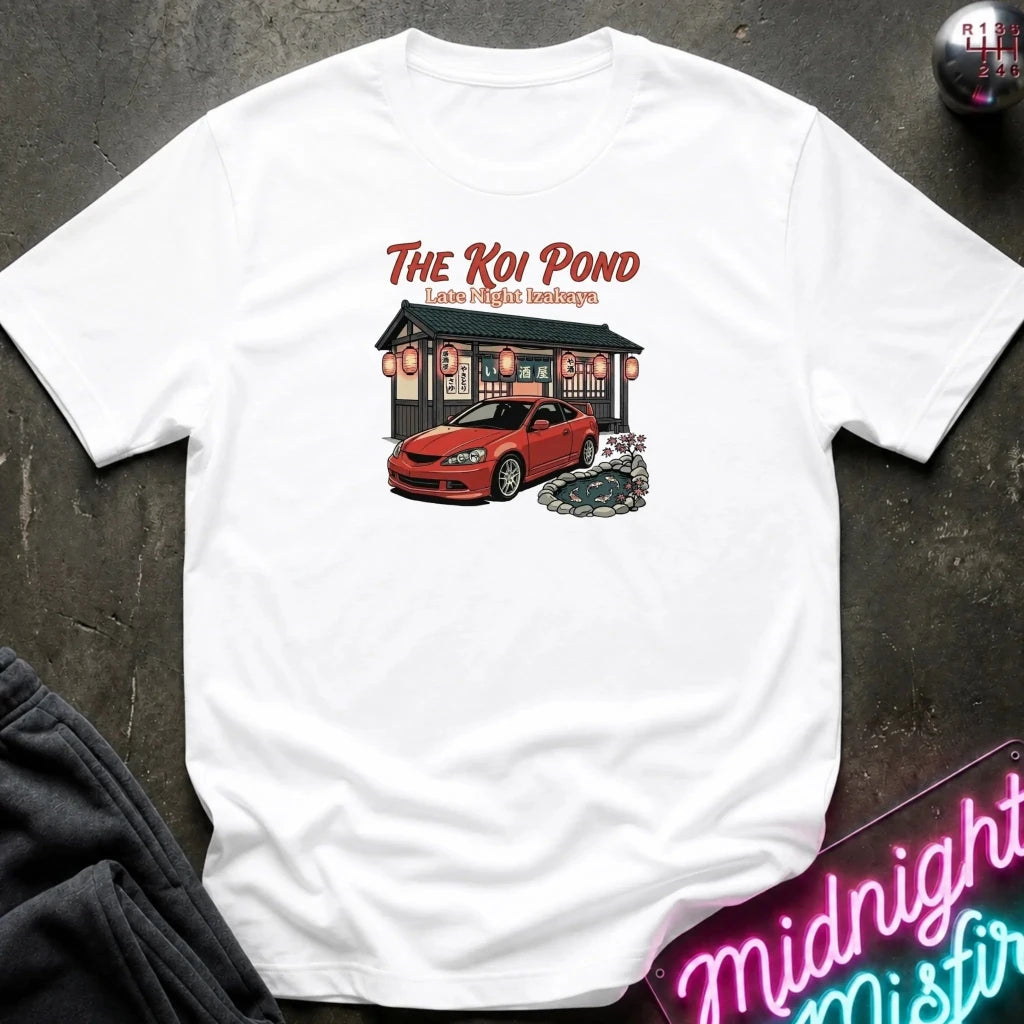 A white short-sleeve graphic t-shirt laid flat on a dark textured surface. The graphic features the text 'THE KOI POND Late Night Hamburger' above an illustration of a red sports car parked in front of a restaurant with red lanterns and a koi pond.