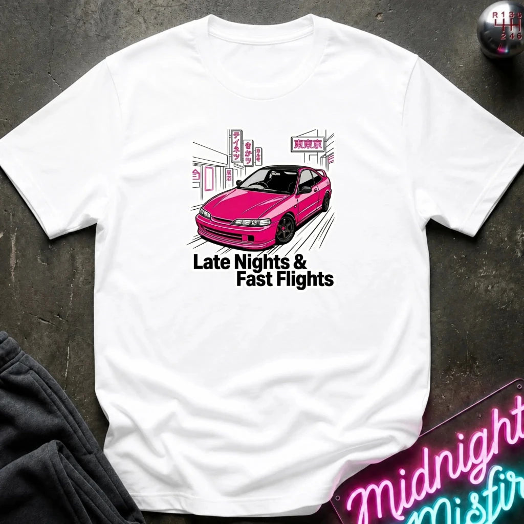 A white short-sleeve graphic T-shirt laid flat on a dark textured surface. The shirt features a graphic of a pink car in front of a city street scene with the text 'Late Nights & Fast Flights' printed below.