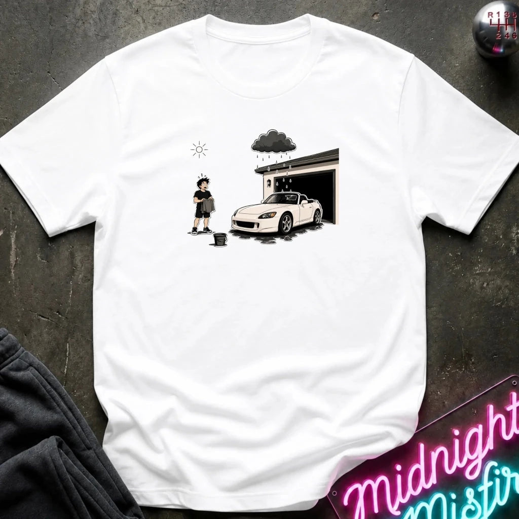A white short-sleeve t-shirt laid flat on a dark textured surface, featuring a black and white graphic print of a person washing a white sports car outside a garage under a dark cloud.