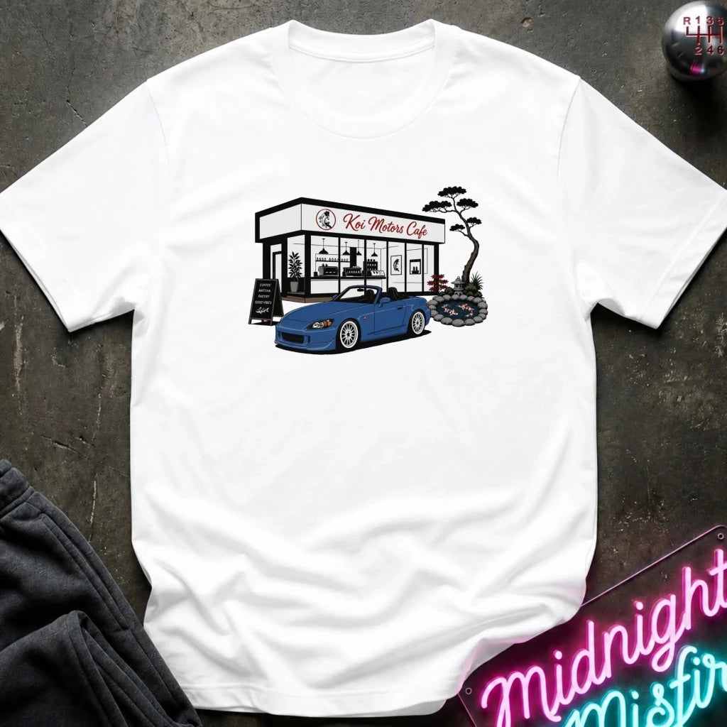 A white short-sleeve t-shirt laid flat on a dark textured surface, featuring a graphic print of a blue sports car parked in front of 'Koi Motors Cafe' with a small tree illustration. 