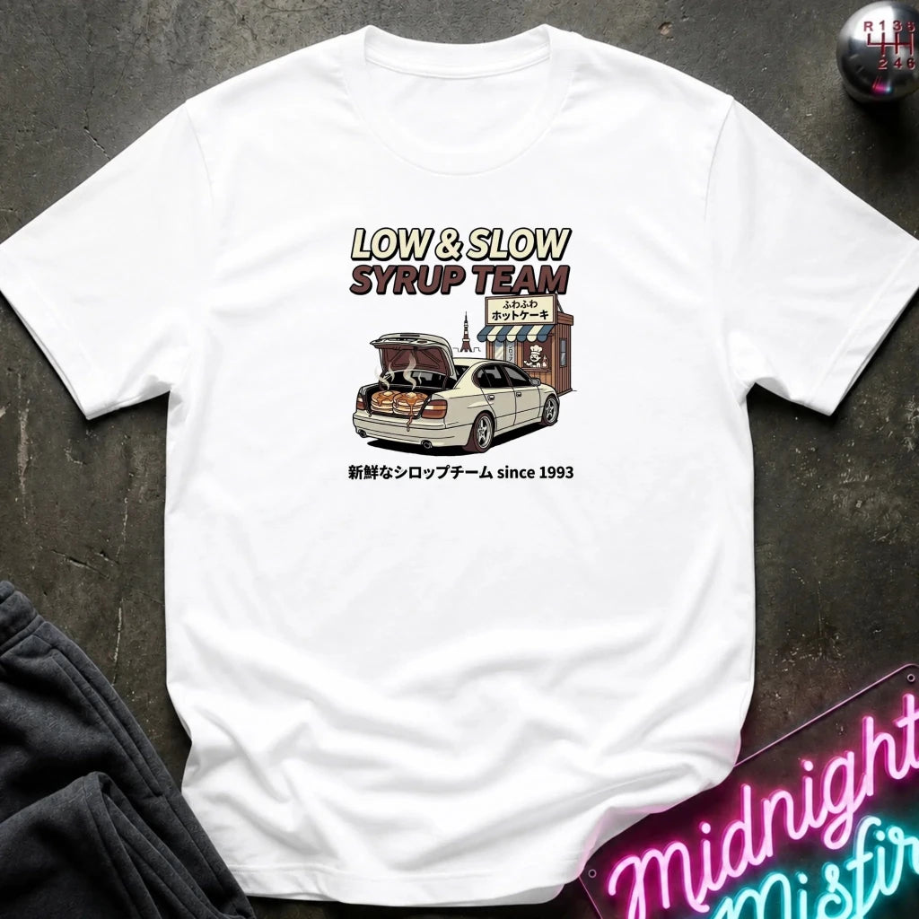 A white short-sleeve graphic t-shirt laid flat on a dark textured surface. The graphic features the text 'LOW & SLOW SYRUP TEAM' above an illustration of a white sedan with its trunk open, parked in front of a shop, with Japanese text below.