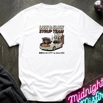 A white short-sleeve graphic t-shirt laid flat on a dark textured surface. The graphic features the text 'LOW & SLOW SYRUP TEAM' above an illustration of a white sedan with its trunk open, parked in front of a shop, with Japanese text below.