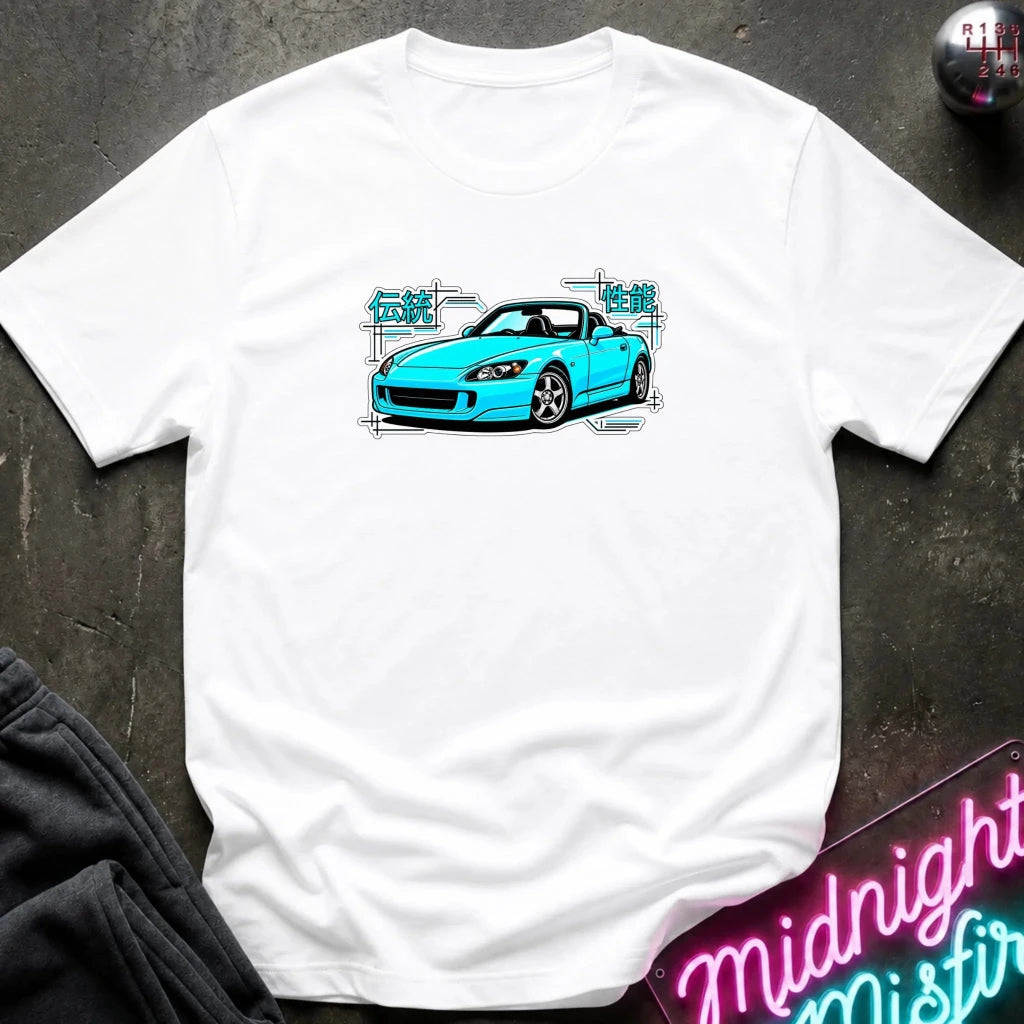 A white t-shirt laid flat on a dark textured surface, featuring a graphic print of a bright blue sports car with white and teal text elements. A neon sign and dark clothing are partially visible in the styled flat-lay composition.