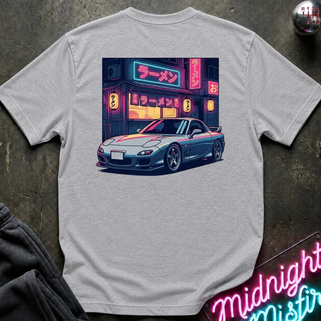 A light gray heather crewneck t-shirt featuring a colorful graphic print of a blue sports car parked in front of a ramen shop with neon signs, laid flat on a dark textured surface.