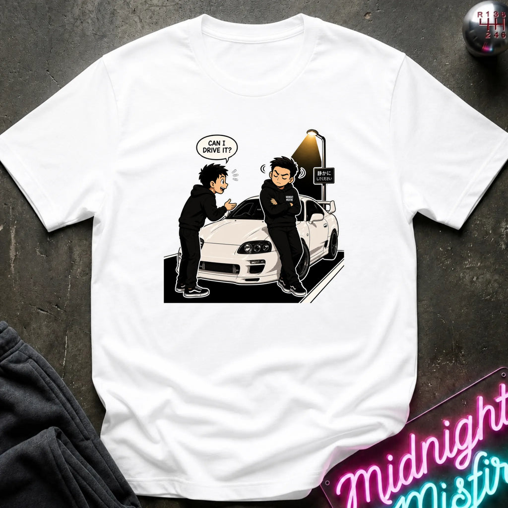 A white short-sleeve graphic t-shirt laid flat on a dark textured surface. The graphic shows a cartoon scene of two people standing next to a white sports car, with one person wearing a black hoodie and the other having a speech bubble that says 'CAN I DRIVE IT?'.