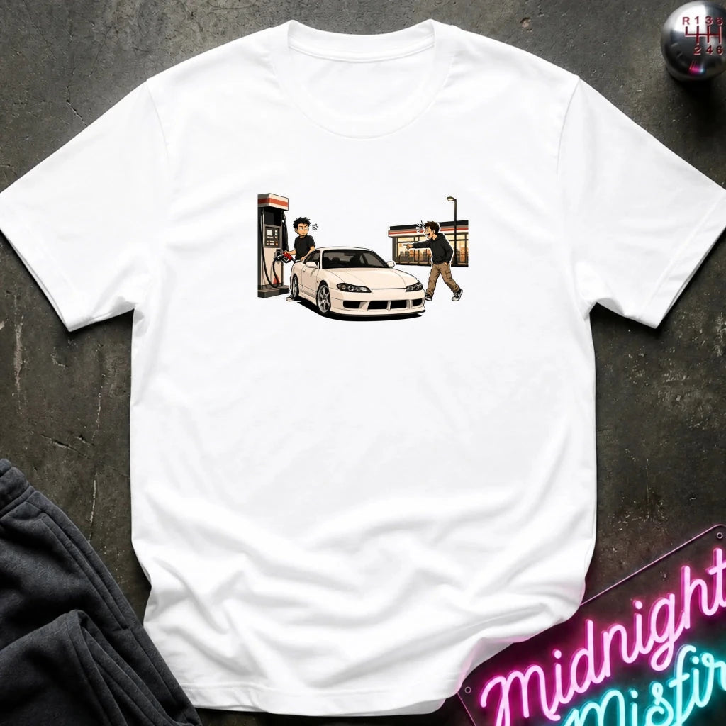 A white short-sleeve crewneck t-shirt laid flat on a dark textured surface, featuring a graphic print of a white sports car at a gas station with two characters. 