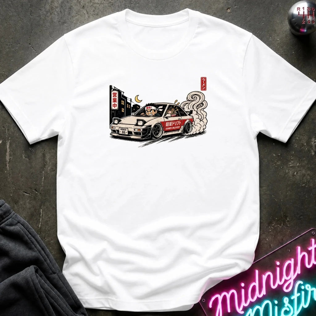A white short-sleeve t-shirt with a graphic print of a drifting car, laid flat on a dark textured surface with a neon sign and dark pants partially visible.