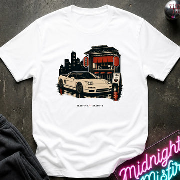 A white short-sleeve crewneck t-shirt featuring a graphic print of a beige sports car parked in front of a Japanese-style building with red lanterns, displayed on a dark textured surface with a neon sign and dark pants partially visible.