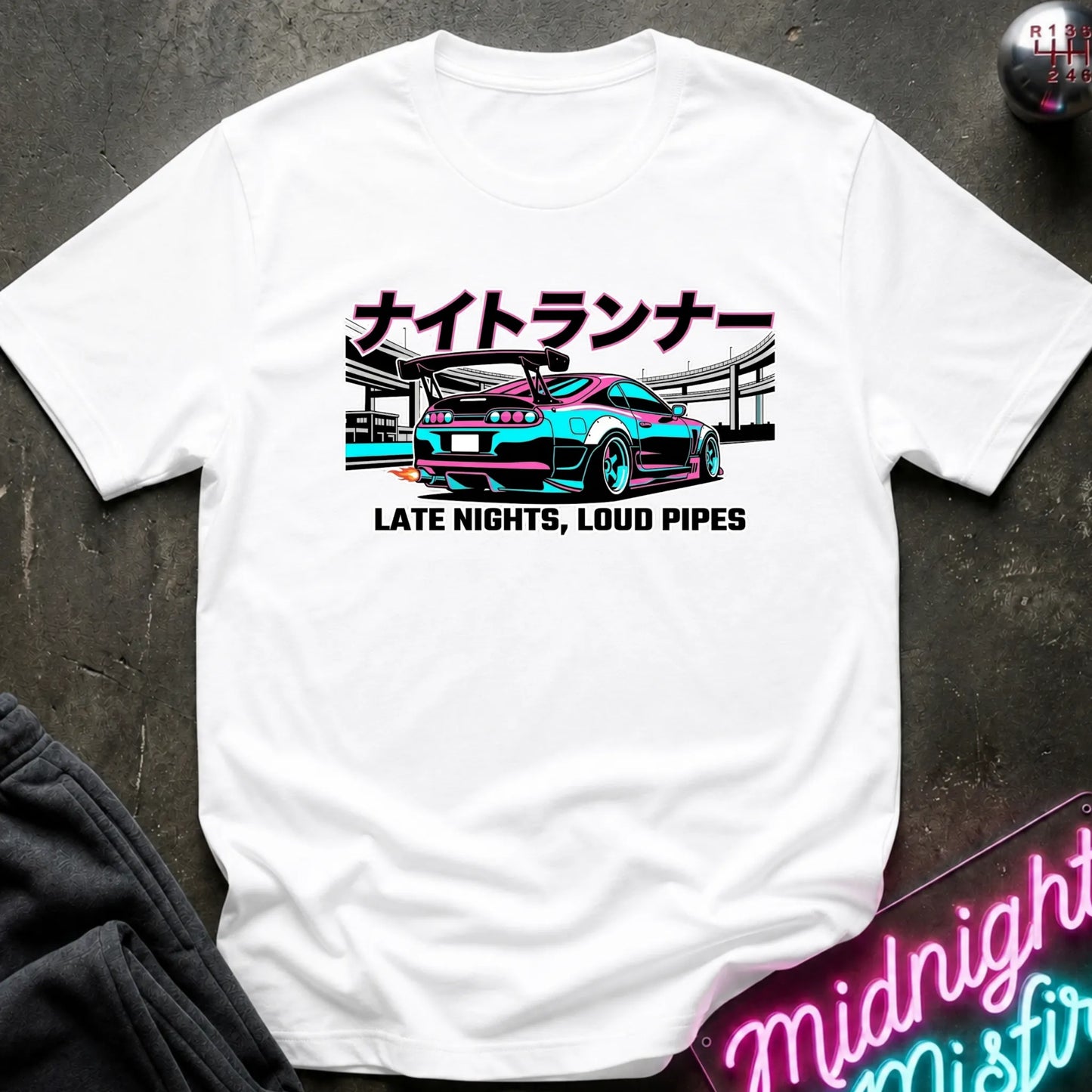 A white short-sleeve graphic T-shirt laid flat on a dark textured surface. The shirt features a colorful print of a sports car with Japanese text above and the phrase 'LATE NIGHTS, LOUD PIPES' below.