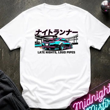 A white short-sleeve graphic T-shirt laid flat on a dark textured surface. The shirt features a colorful print of a sports car with Japanese text above and the phrase 'LATE NIGHTS, LOUD PIPES' below.