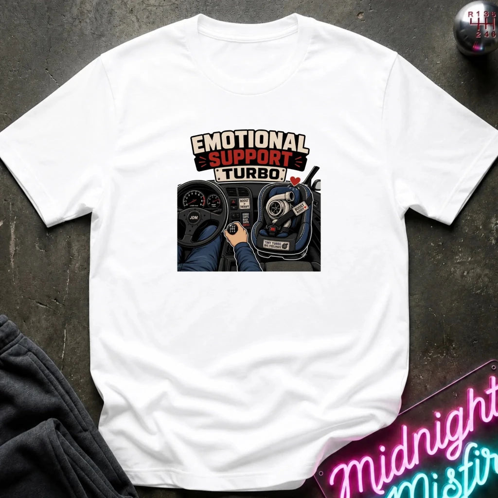 A white short-sleeve graphic T-shirt laid flat on a dark textured surface. The shirt features a central graphic with the text 'EMOTIONAL SUPPORT TURBO' above an illustration of a car interior with a hand on the steering wheel.