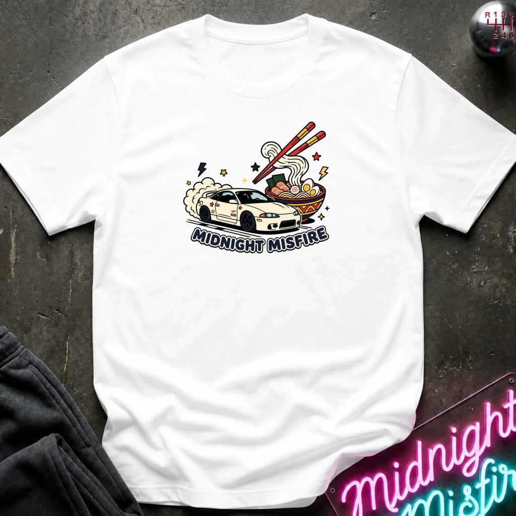 A white short-sleeve graphic T-shirt laid flat on a dark textured surface. The shirt features a colorful illustration of a white sports car drifting next to a bowl of ramen with chopsticks, with the text 'MIDNIGHT MISFIRE' printed below the graphic.