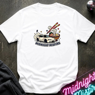 A white short-sleeve graphic T-shirt laid flat on a dark textured surface. The shirt features a colorful illustration of a white sports car drifting next to a bowl of ramen with chopsticks, with the text 'MIDNIGHT MISFIRE' printed below the graphic.