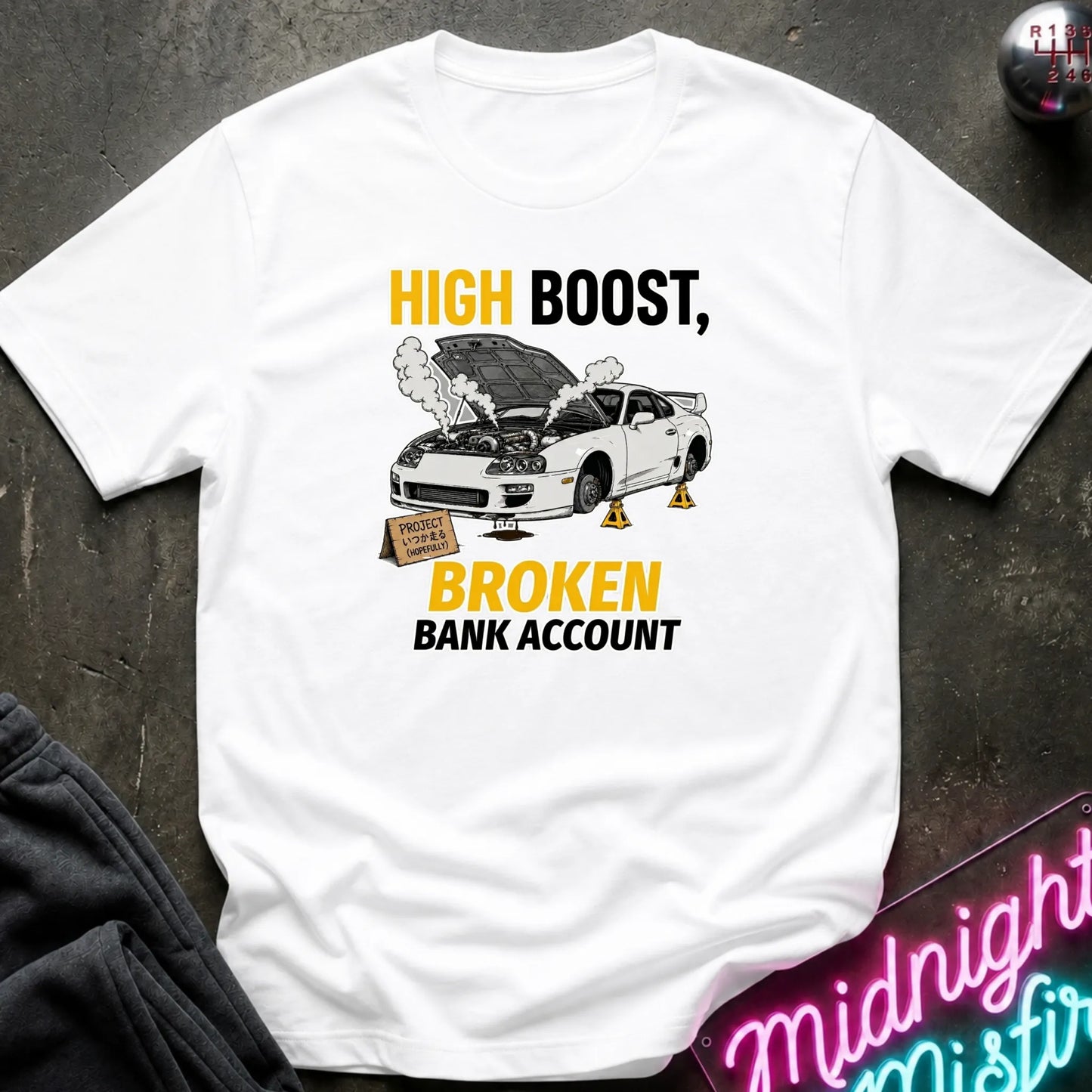 A white short-sleeve graphic T-shirt laid flat on a dark textured surface. The shirt features a printed design with the text 'HIGH BOOST, BROKEN BANK ACCOUNT' in yellow and black, alongside an illustration of a white sports car with its hood open, smoke, and yellow jack props.