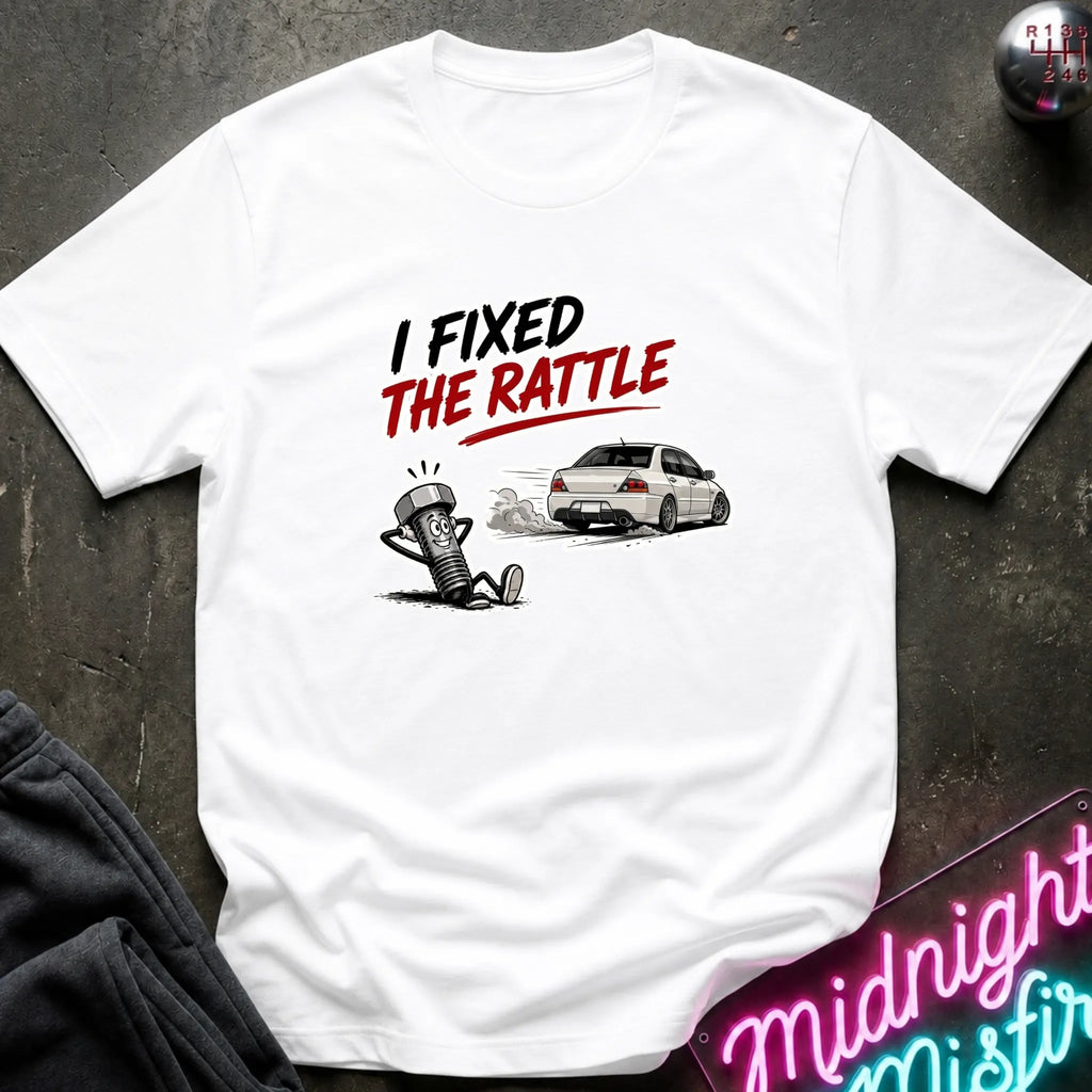 A white short-sleeve graphic T-shirt laid flat on a dark textured surface. The shirt features a printed design with the text 'I FIXED THE RATTLE' in black and red, above a cartoon character and a white sports car.