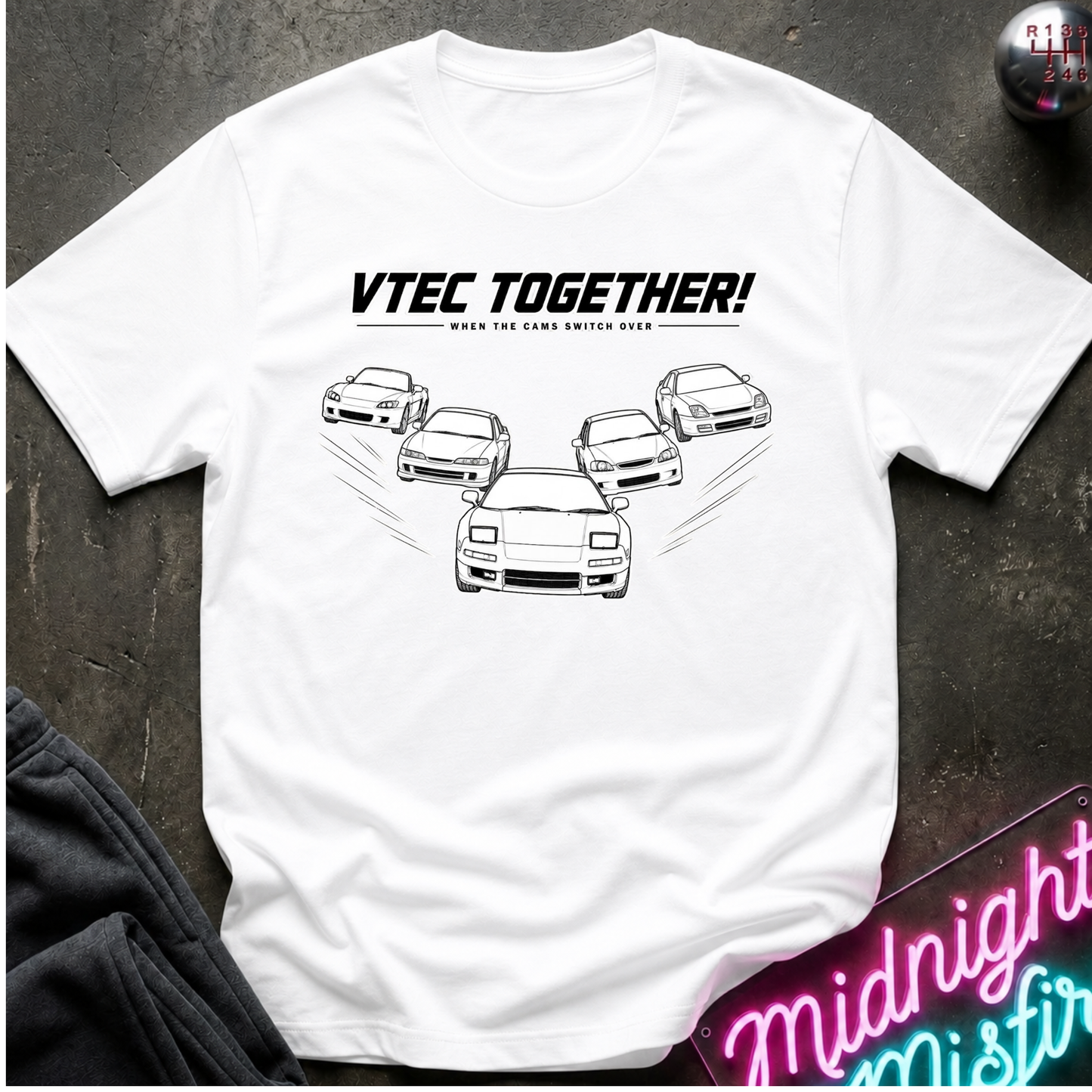 A white short-sleeve graphic T-shirt laid flat on a dark textured surface. The shirt features the text 'VTEC TOGETHER!' with smaller text underneath and a line drawing of several sports cars arranged in a V formation.