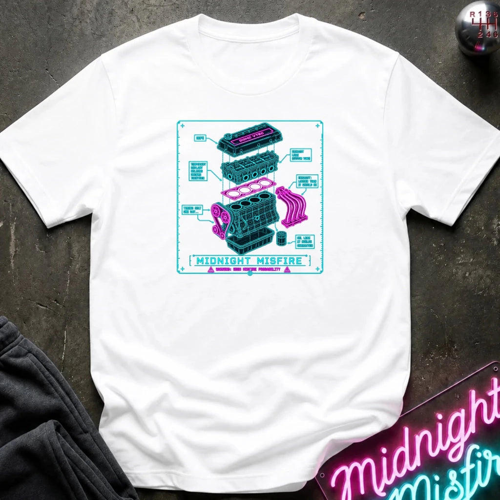 white short-sleeve graphic T-shirt laid flat on a dark textured surface. The shirt features a centered rectangular print with a light blue and pink exploded-style engine diagram and the text 'MIDNIGHT MISFIRE.