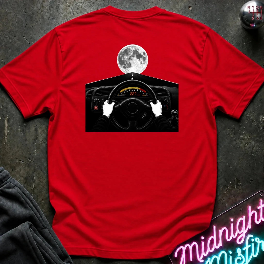A red short-sleeve t-shirt laid flat on a dark textured surface, featuring a graphic print of hands on a car steering wheel with a full moon above. A gear shift knob and a neon sign are partially visible as props.