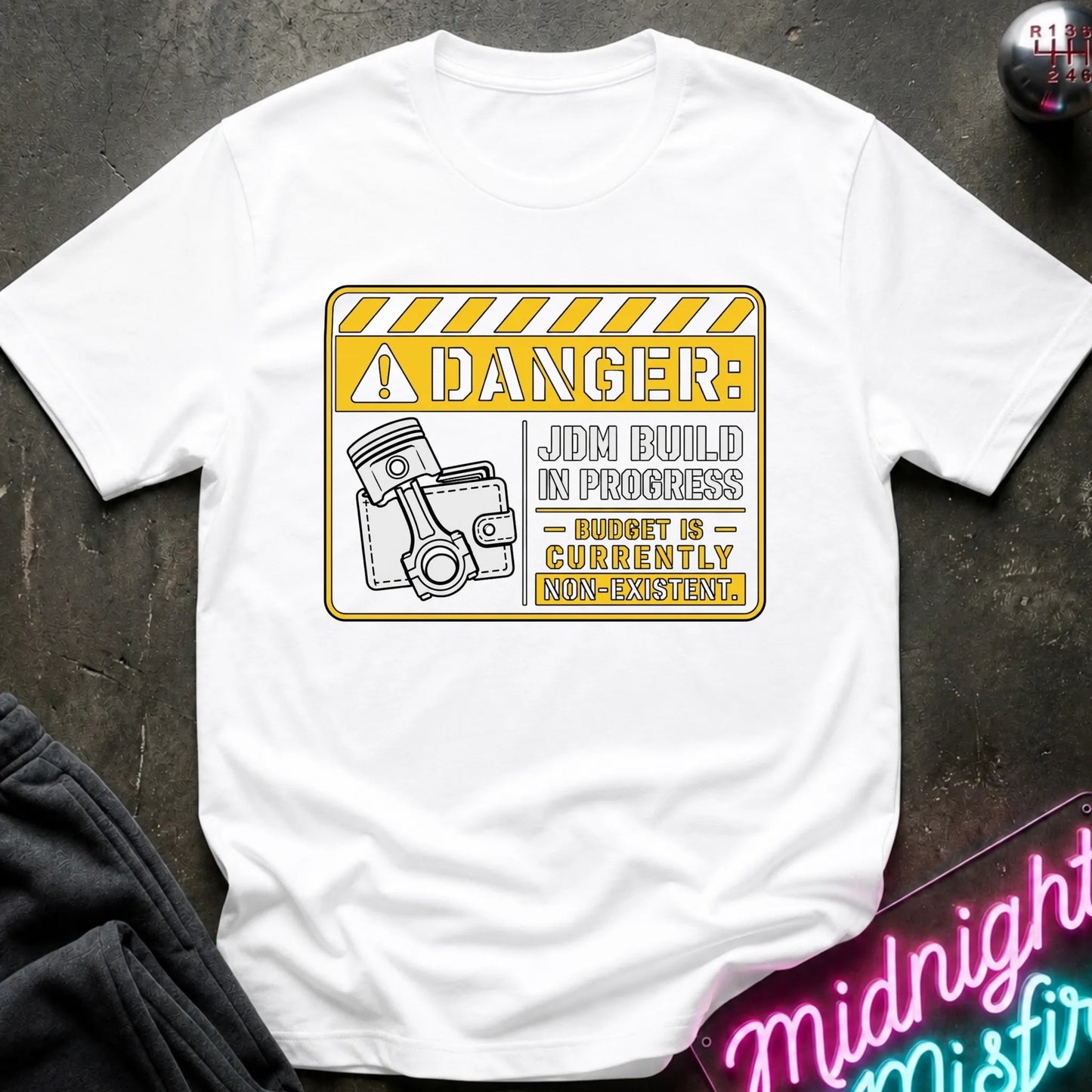 A white short-sleeve graphic T-shirt laid flat on a dark textured surface. The shirt features a yellow and white warning-sign style print with the text 'DANGER: JDM BUILD IN PROGRESS - BUDGET IS - CURRENTLY NON-EXISTENT'.