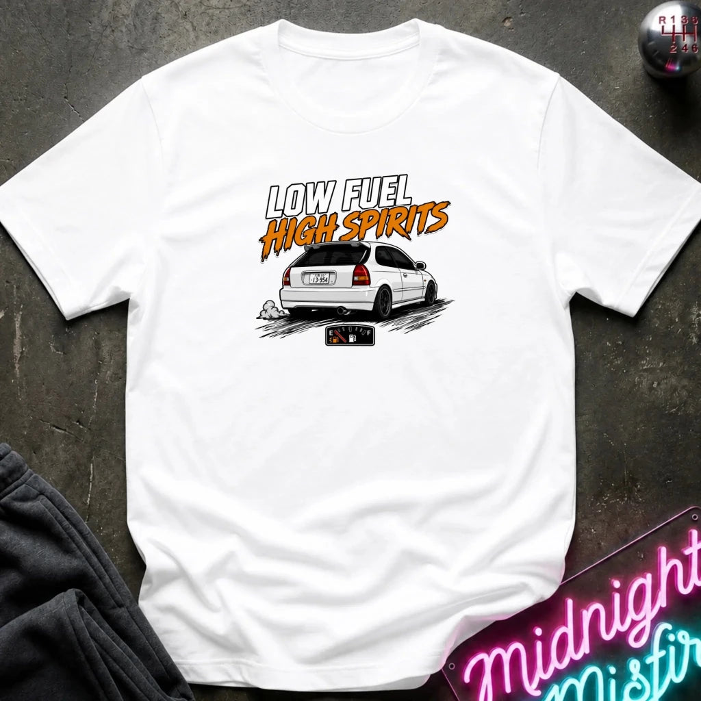 A white short-sleeve graphic T-shirt laid flat on a dark textured surface. The shirt features a printed design of a white hatchback car with the text 'LOW FUEL HIGH SPIRITS' in white and orange lettering.