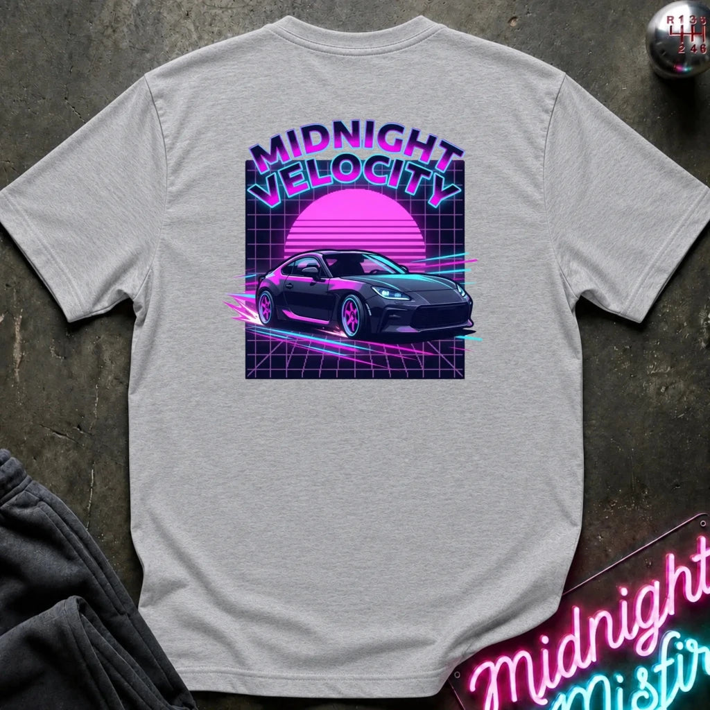 A light gray heather t-shirt laid flat on a dark textured surface, featuring a colorful retro 80s graphic on the back with the text 'MIDNIGHT VELOCITY', a pink sun, and a black sports car.