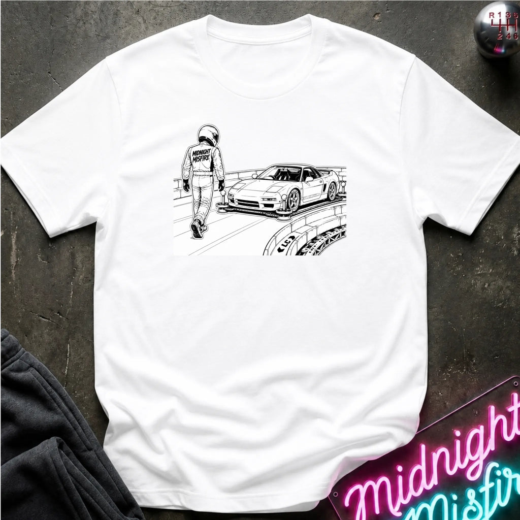 A white short-sleeve crewneck t-shirt laid flat on a dark textured surface, featuring a black line art graphic of a race car and a driver on the chest.