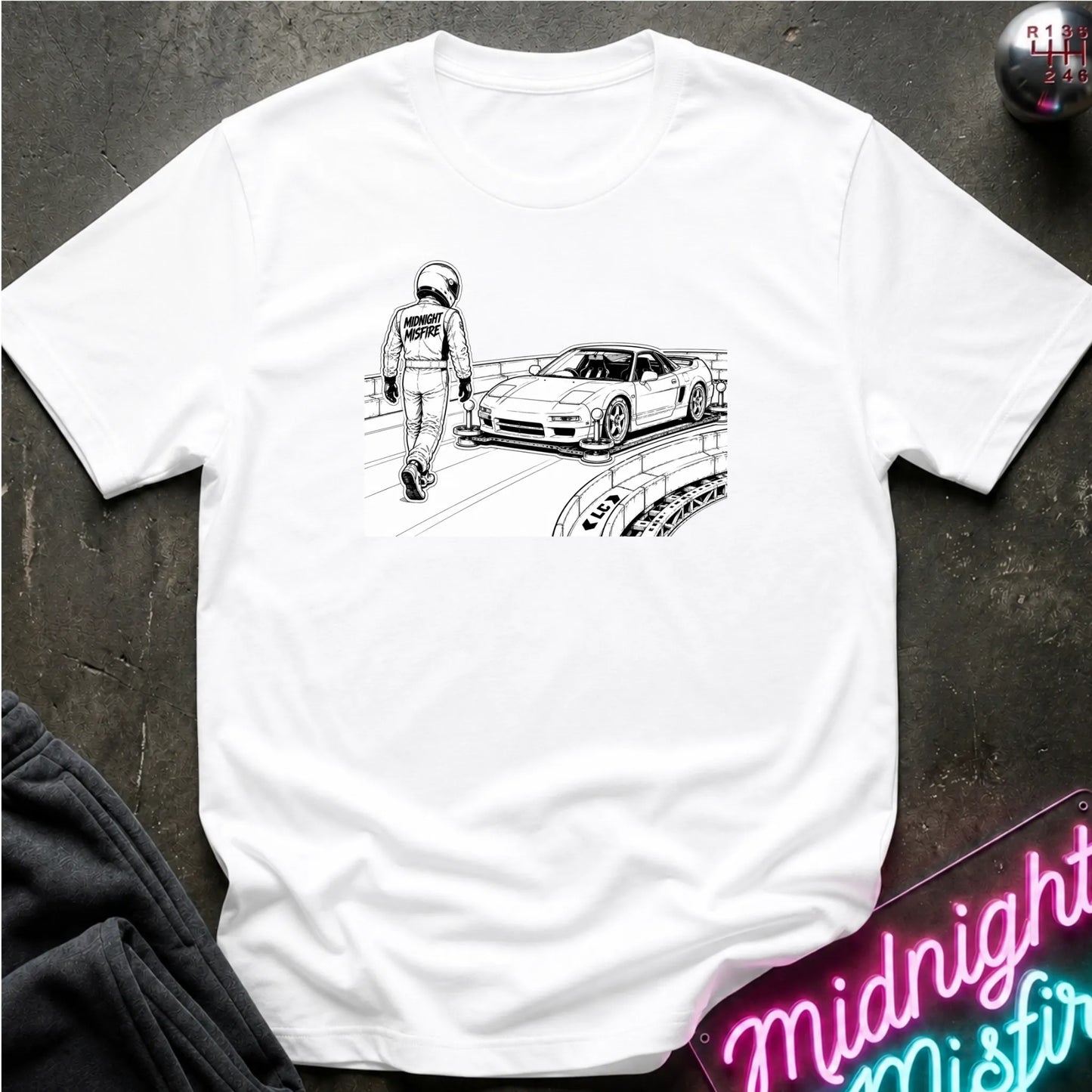 A white short-sleeve crewneck t-shirt laid flat on a dark textured surface, featuring a black line art graphic of a race car and a driver on the chest.