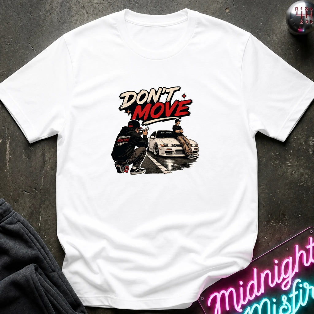 A white short-sleeve graphic t-shirt laid flat on a dark textured surface. The graphic features the words 'DON'T MOVE' in a stylized red and black font above an illustration of a person crouching next to a white sports car.