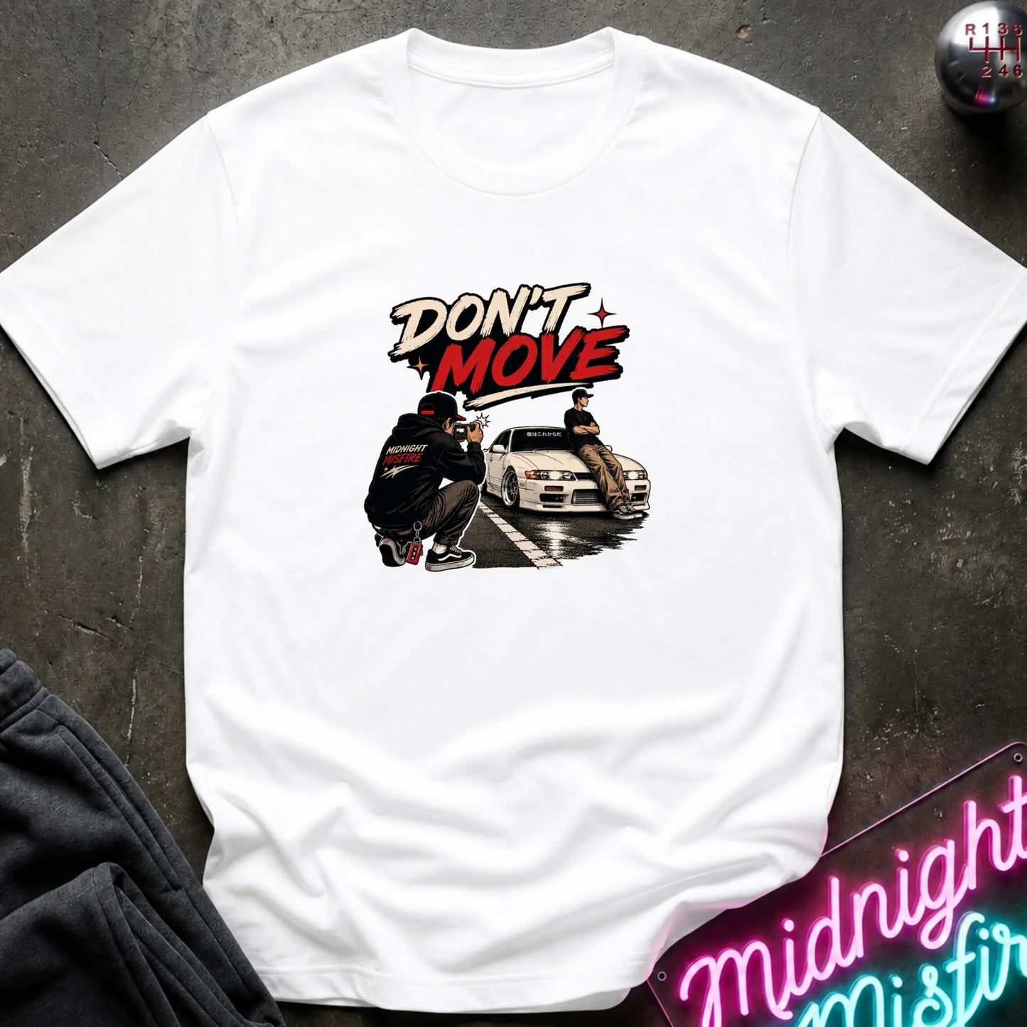 A white short-sleeve graphic t-shirt laid flat on a dark textured surface. The graphic features the words 'DON'T MOVE' in a stylized red and black font above an illustration of a person crouching next to a white sports car.