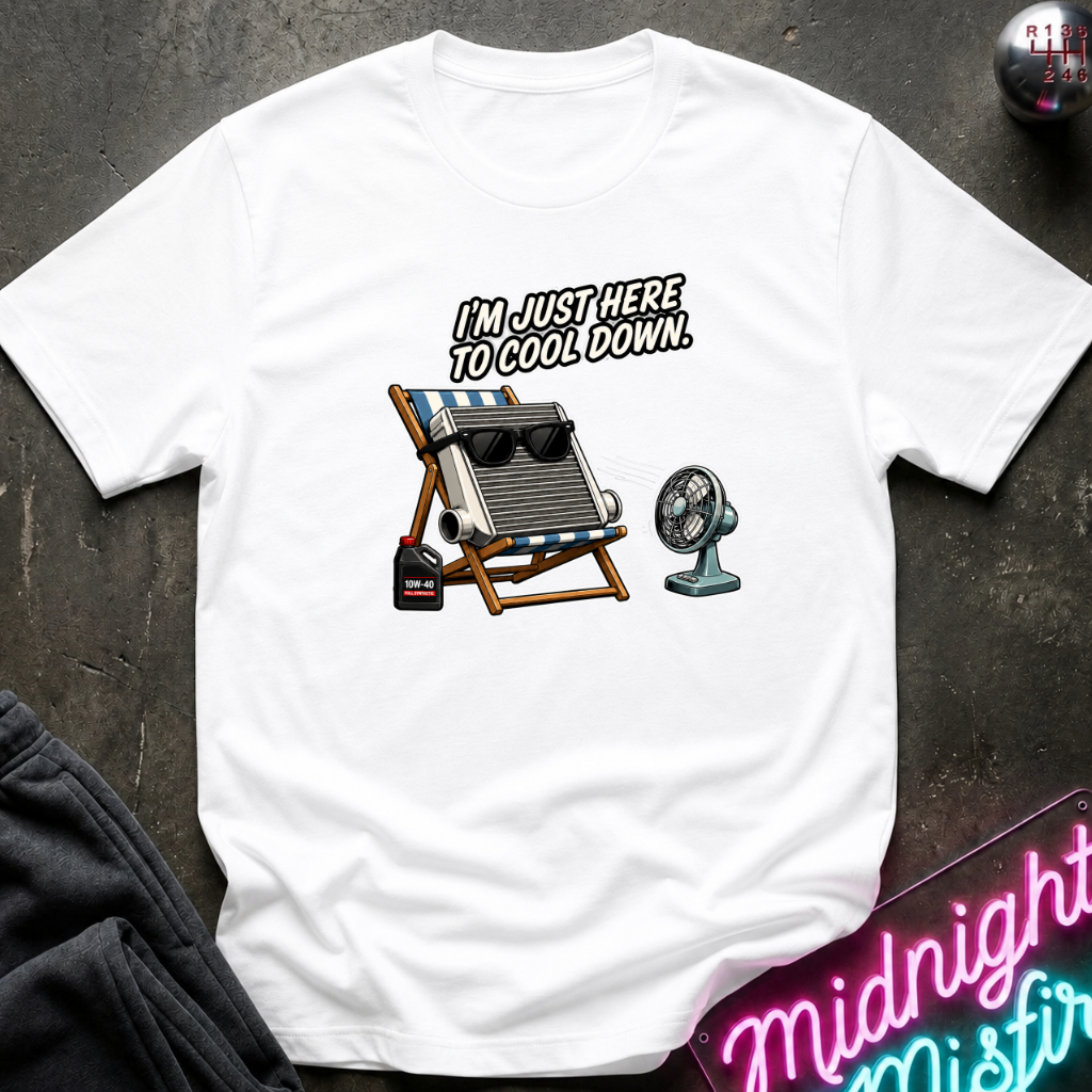 A white short-sleeve graphic t-shirt laid flat on a dark textured surface. The graphic features a white car radiator sitting in a striped beach chair next to a small fan, with the text 'I'M JUST HERE TO COOL DOWN.