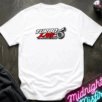 A white short-sleeve t-shirt laid flat on a dark textured surface, featuring a 'Turbo Lag' car logo graphic in red, black, and white on the chest. Styled flat-lay props include a dark sweatshirt in the bottom-left corner and a pink neon sign in the bottom-right corner.