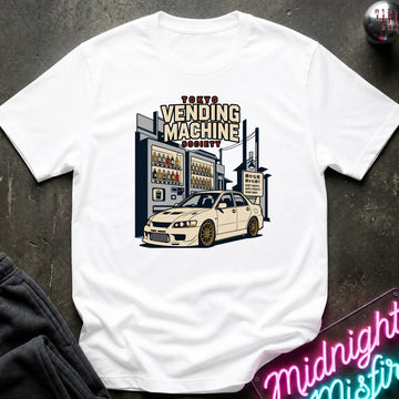 A white short-sleeve graphic t-shirt laid flat on a dark textured surface. The graphic features a vintage-style illustration of a white sports car parked in front of a vending machine building, with the text 'TOKYO VENDING MACHINE SOCIETY' above it.