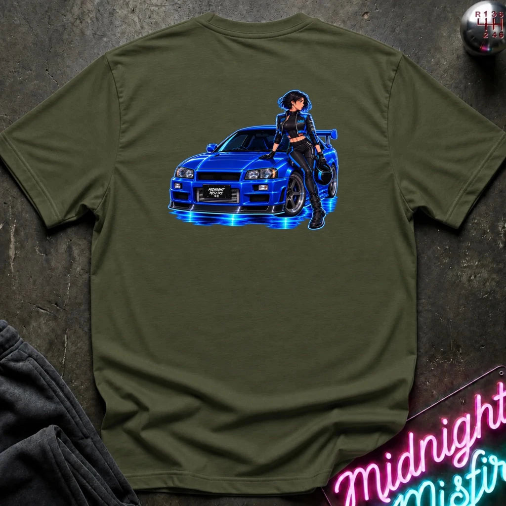 An olive green short-sleeve t-shirt laid flat on a dark textured surface, featuring a graphic print of a blue sports car with glowing blue neon lights and an anime-style woman in a black outfit. A neon sign and folded dark fabric are partially visible in the corners.