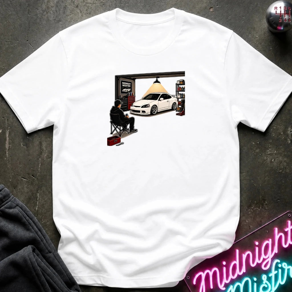 A white short-sleeve t-shirt laid flat on a dark textured surface, featuring a graphic print of a person sitting in a chair in front of a garage with a white sports car inside. A neon sign reading 'Midnight Mistral' and dark gray sweatpants are partially visible as props.