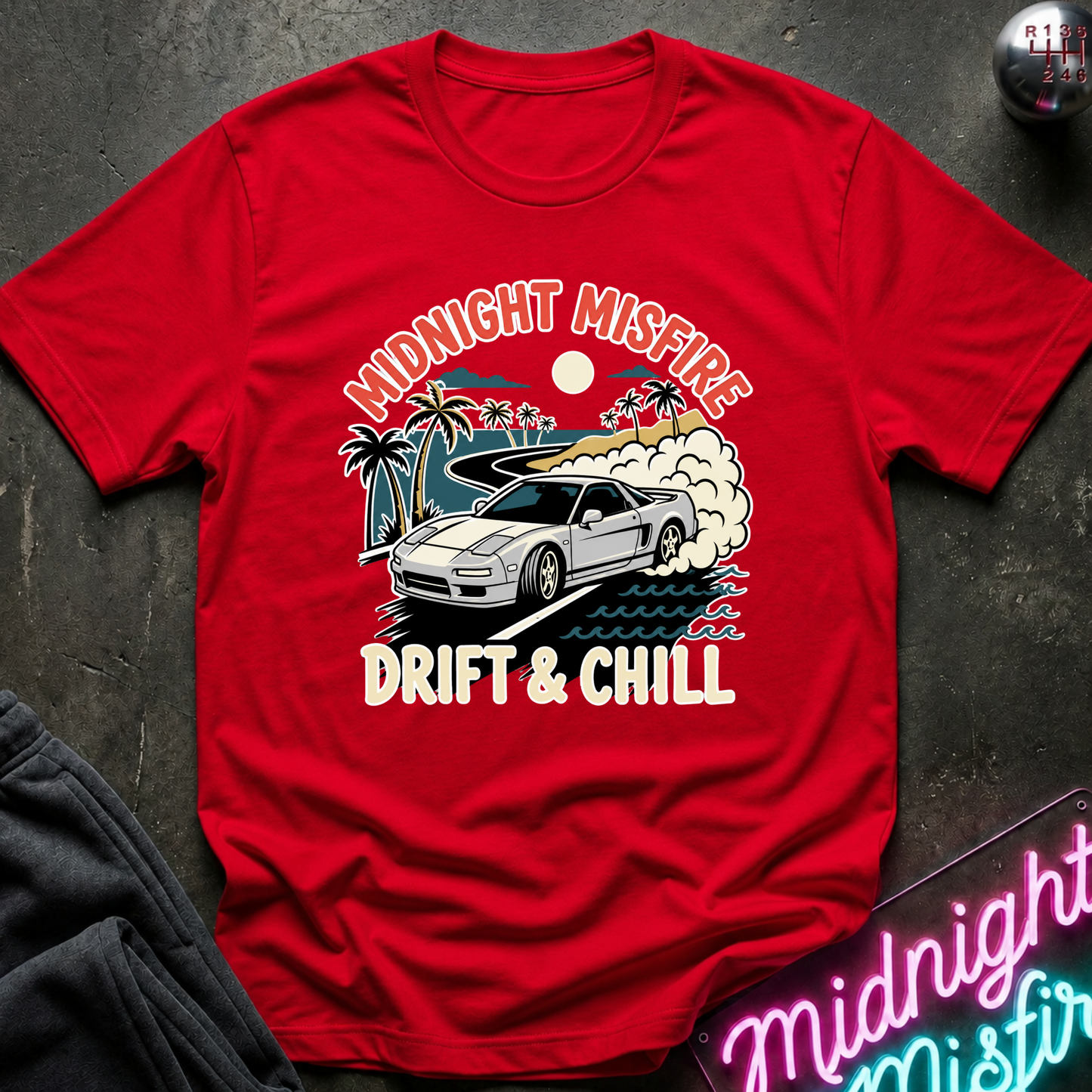 A red short-sleeve graphic T-shirt laid flat on a dark textured surface. The shirt features a retro-style print of a silver sports car drifting on a road with palm trees, smoke, and the text 'MIDNIGHT MISFIRE' above and 'DRIFT & CHILL' below.