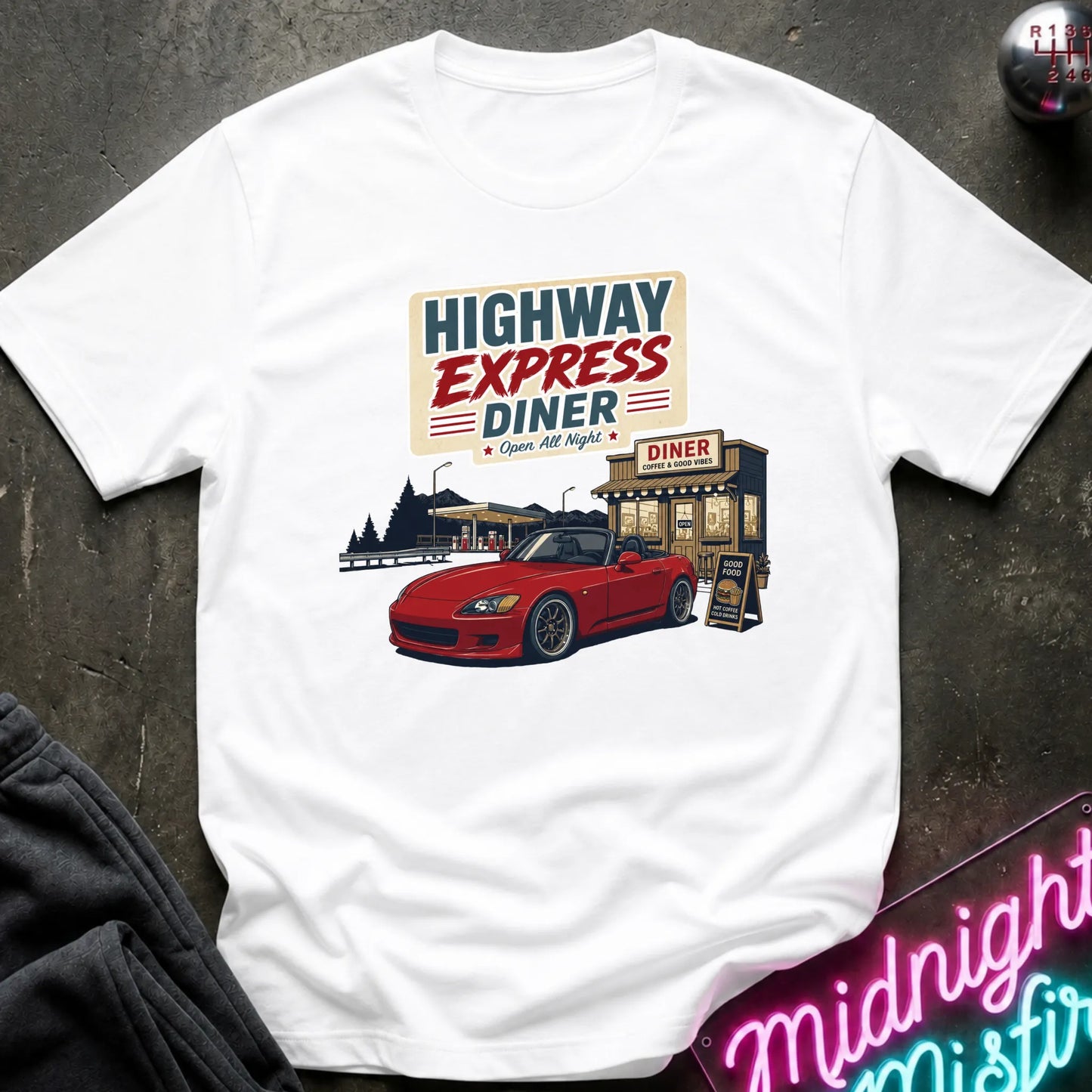 A white short-sleeve graphic T-shirt laid flat on a dark textured surface. The shirt features a retro-style print reading 'HIGHWAY EXPRESS DINER' above an illustration of a red convertible car parked outside a diner.